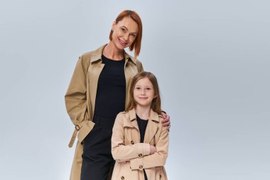 two generations, woman with red hair and little girl standing in trench coats on grey background clipart