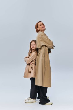 two generations, happy redhead woman and girl standing in trench coats with folded arms in studio clipart