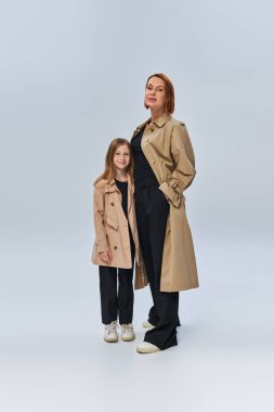 two female generations, redhead woman and happy girl standing in trench coats on grey background clipart