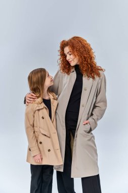 two female generations, redhead woman looking at daughter and standing in coats on grey background clipart