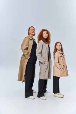joyful redhead family in coats posing with hands in pockets on grey background, female generations clipart