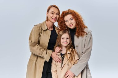 redhead women in autumn coats hugging little girl on grey background, three female generations clipart
