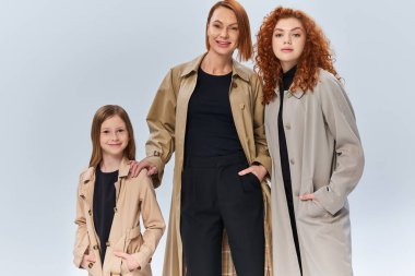 Gri arka planda elleri ceplerinde poz veren sonbahar ceketli kızıl saçlı aile, kadın nesiller.