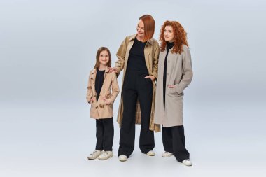 redhead family in autumn coats posing with hands in pockets on grey backdrop, three generations clipart