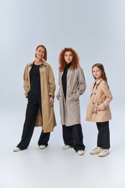 Gri arka planda elleri ceplerinde poz veren şık paltolu kızıl saçlı bir aile, üç kuşak.