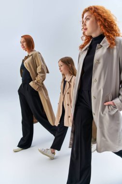 three generation redhead family walking with hands in pockets of trench coats on grey backdrop clipart