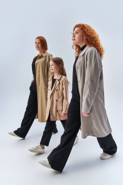 three generation redhead family walking with hands in pockets of stylish coats on grey backdrop clipart