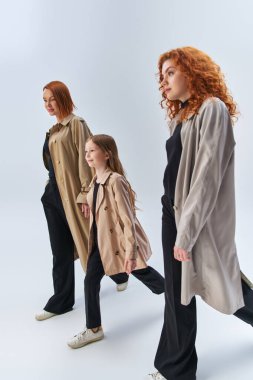three generation redhead family walking together in stylish coats on grey backdrop, women and girl clipart