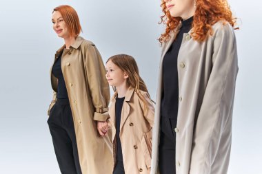 Üç kuşak kızıl saçlı aile, sonbahar modasında gri arka planda şık paltolarla birlikte yürüyorlar.