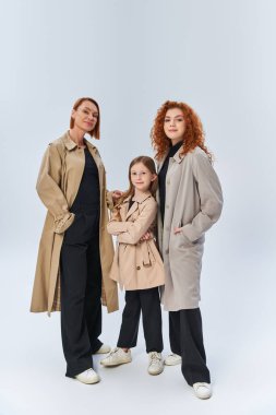 Üç kuşak neşeli aile, gri arka planda şık paltolarla, sonbahar modasında bir arada duruyorlar.