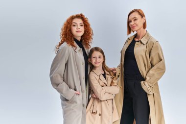 three generation joyful family standing together in stylish coats on grey backdrop, autumn fashion clipart