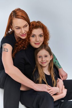 Üç kuşak konsepti, eşleşen kıyafetler içindeki pozitif kızıl saçlı aile gri arka planda sarılıyor.