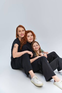 Gri arka planda birbirine uyumlu kıyafetlerle sarılıp oturan kızıl saçlı bir aile. Üç nesildir.