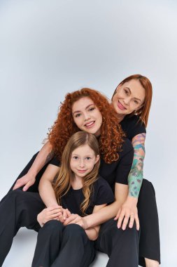 Kızıl saçlı mutlu bir aile, gri arka planda aynı kıyafetlerle oturuyor, üç nesildir.