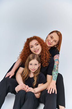happy family with red hair sitting in matching outfits on grey backdrop, three generations clipart