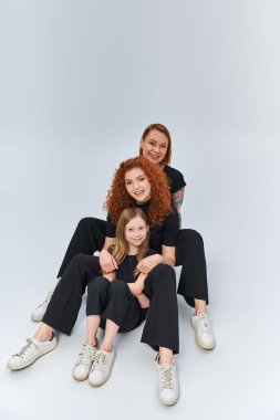 Gri arka planda birbirine uyumlu kıyafetlerle sarılıp oturan kızıl saçlı bir aile. Üç nesildir.