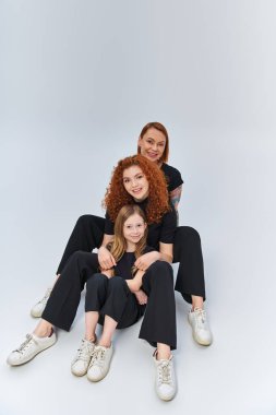 redhead family in matching outfits hugging and sitting together on grey backdrop, three generations clipart