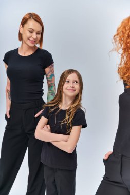 Gri arka planda eşleşen kıyafetler giymiş kadınların yanında duran kızıl saçlı kız. Nesiller boyu.