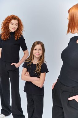 happy girl standing with folded arms near redhead family in matching clothes on grey backdrop clipart