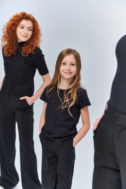 Neşeli kız kollarını kavuşturmuş, gri arka planda eşleşen kıyafetlerle kızıl saçlı ailenin yanında duruyor.