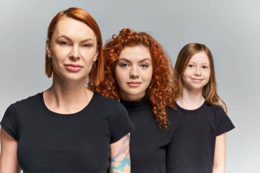 three generations of redhead women and child posing in matching attire on grey backdrop, family clipart