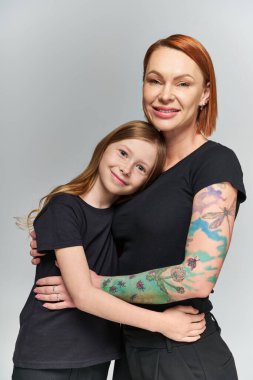 two generations, redhead woman and girl in matching attire hugging on grey backdrop, happiness clipart