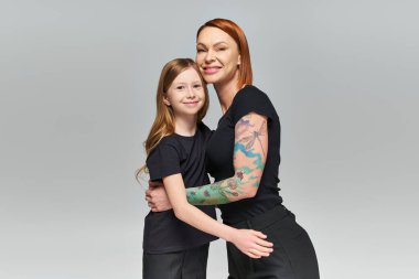 two generations, redhead woman and girl in matching attire hugging on grey backdrop, joyous family clipart
