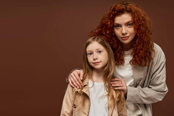 two generations, young curly mother with red hair looking at camera with daughter on brown backdrop
