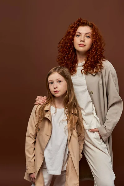 two generations, young curly mother looking at camera with redhead daughter on brown backdrop