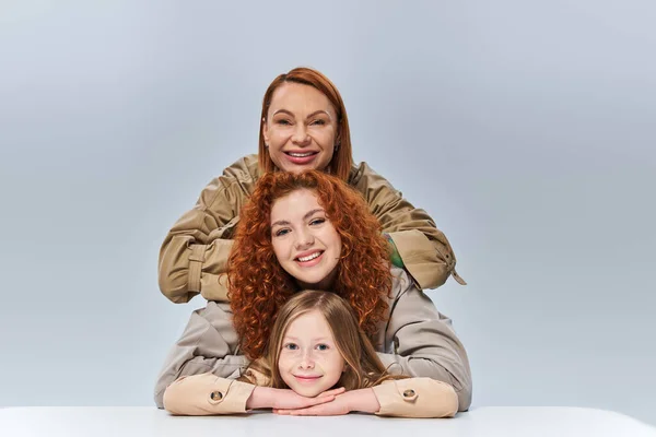 three generations, redhead mother and child in beige trench coats posing on grey background