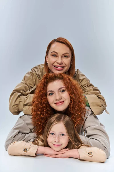 happy three generations, redhead mother and child in beige trench coats posing on grey background