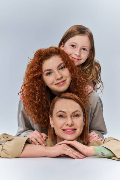 three female generations, happy redhead family in beige coats smiling on grey backdrop, family bond
