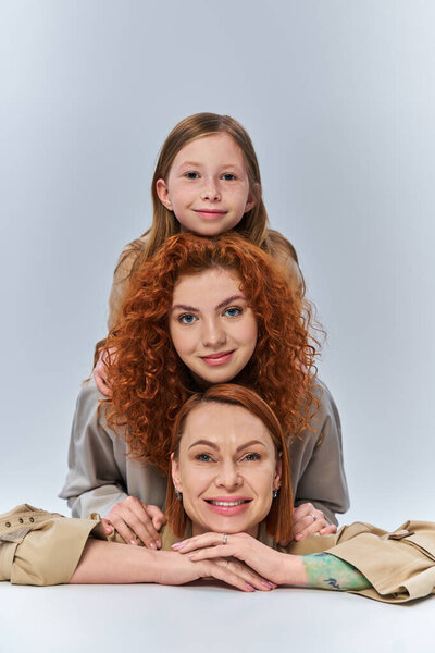 three generations, happy redhead family in beige coats smiling on grey backdrop, female bond