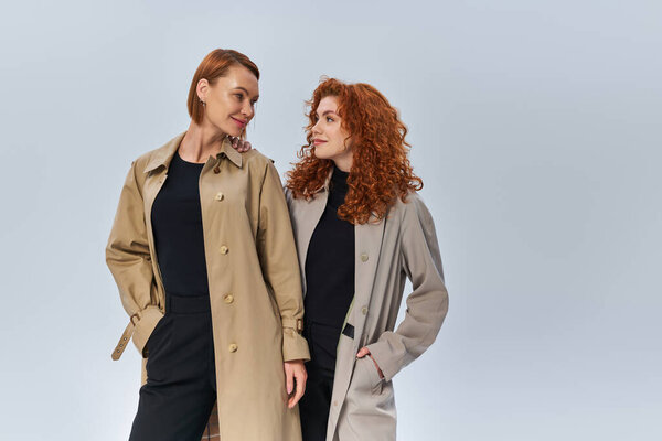 two generations of redhead women posing in coats and looking at each other on grey background