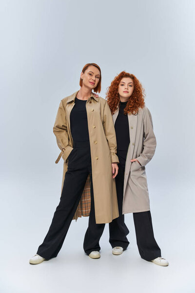 two generations of redhead women posing in autumnal coats on grey background, hands in pockets
