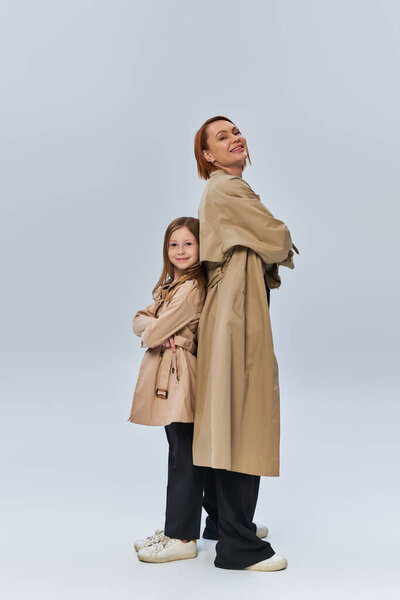 two generations, happy redhead woman and girl standing in trench coats with folded arms in studio