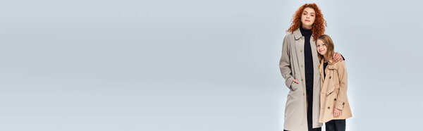 Два поколения, рыжая женщина обнимает дочь и стоя в пальто на сером фоне, баннер