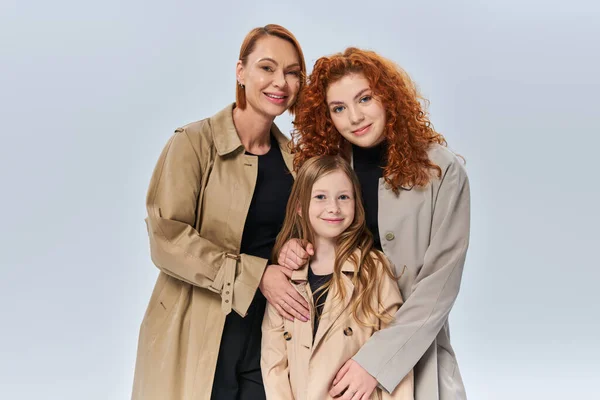 redhead women in autumn coats hugging little girl on grey background, three female generations