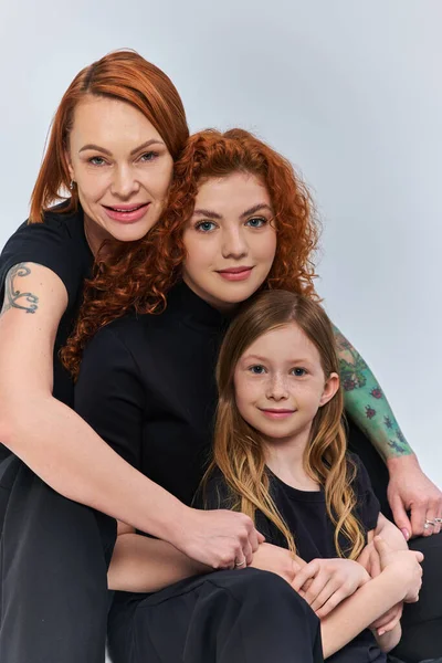 three generations concept, cheerful redhead family in matching outfits hugging on grey backdrop