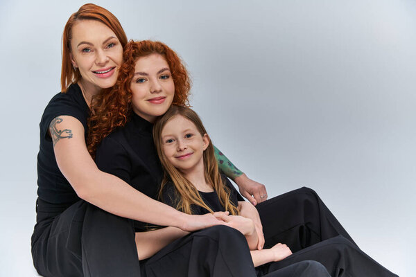 концепция трех поколений, рыжая семья в подходящих нарядах, обнимающая и сидящая на сером фоне
