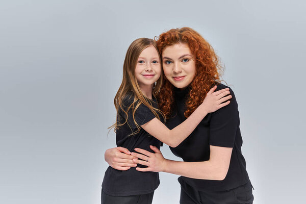 happy young mother with curly red hair hugging little girl on grey backdrop, two generations