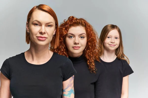 three generations of redhead women and child posing in matching attire on grey backdrop, family