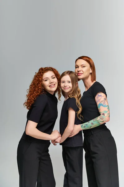 three generations concept, happy redhead women and girl in matching attire hugging on grey backdrop