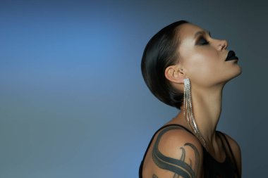 profile of tattooed woman with dark makeup, shiny earring and closed eyes on blue grey backdrop clipart