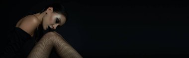 mysterious woman with closed eyes and dark makeup sitting in fishnet tights on black, banner clipart