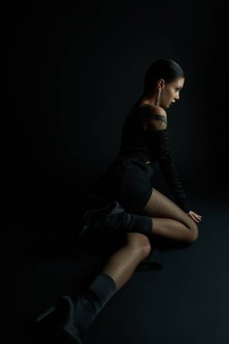 enchanting woman in sexy dress and fishnet tights sitting in darkness and looking away in studio clipart