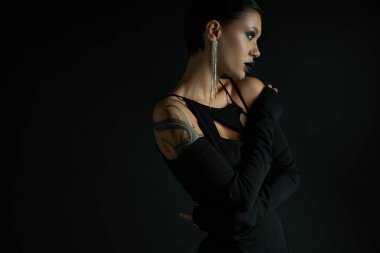 tattooed and glamour woman with eerie makeup posing in sexy dress on black, halloween concept clipart
