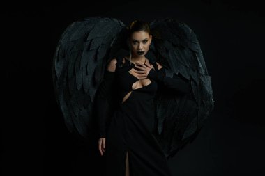 Kara kanatlı düşmüş melek kostümlü seksi kadın Cadılar Bayramı 'nda siyah kameraya bakıyor.