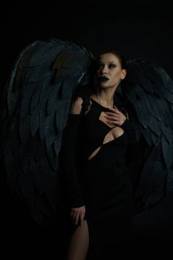tattooed woman in halloween costume of fallen angel with wings looking away on black, demonic charm clipart