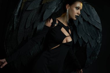 Cadılar bayramı kostümlü büyüleyici bir kadın. Kara melek kanatlarıyla kara arka plana bakıyor.